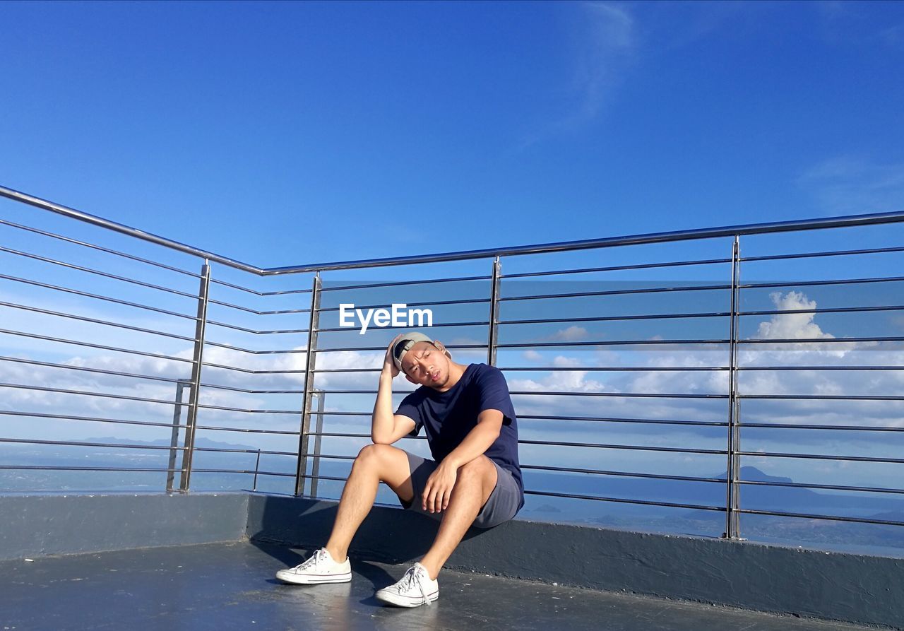 Man sitting on railing against blue sky