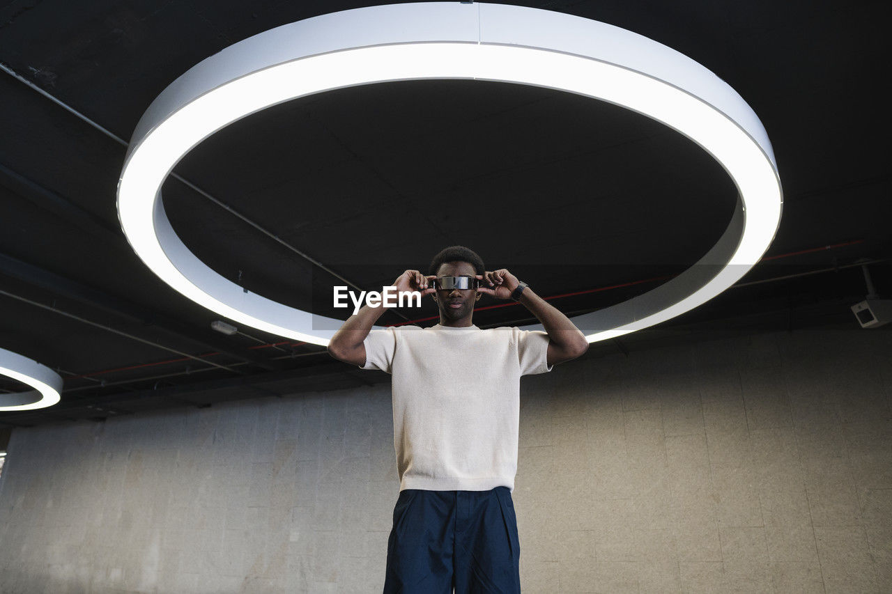 Young man with futuristic cyber glasses standing under modern ring lamp with fingers on temples