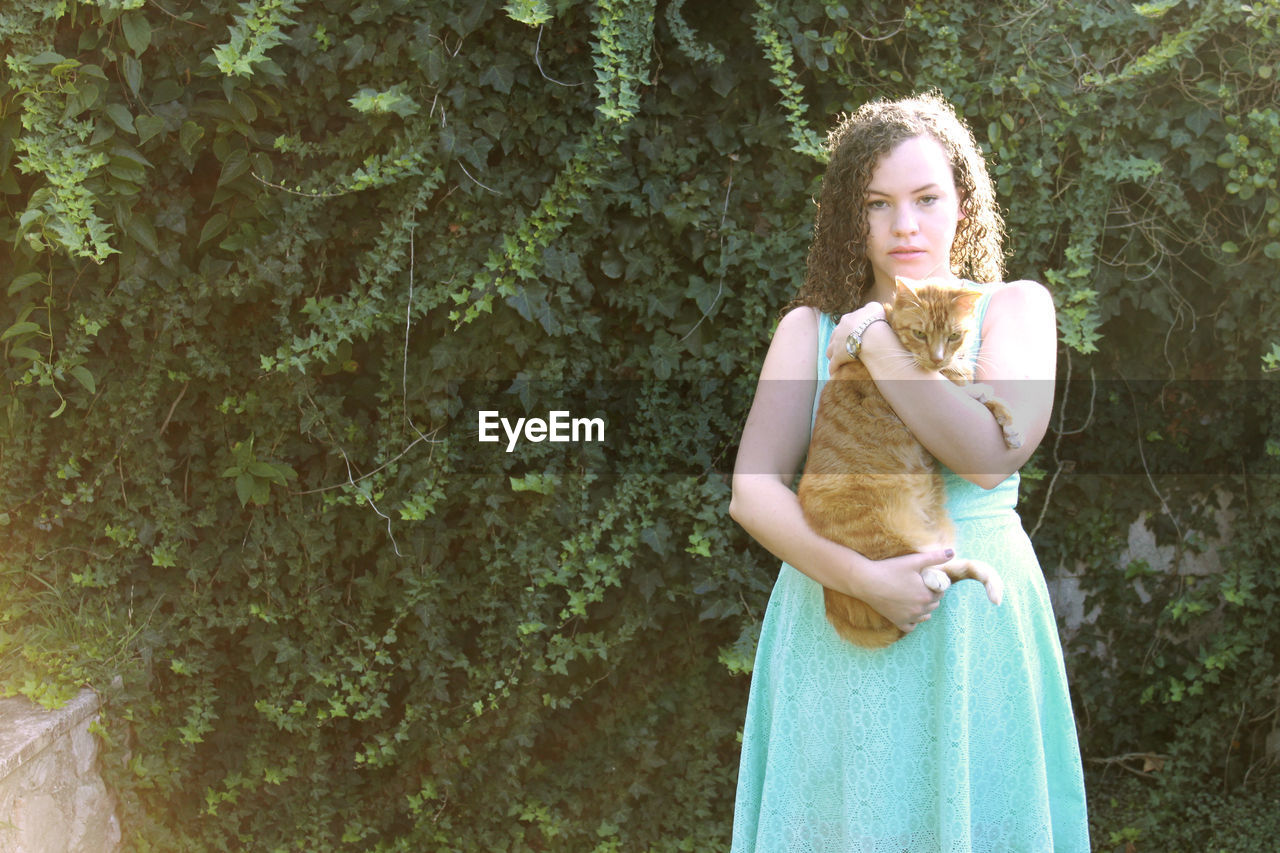 Portrait of young woman holding cat while standing against plants in park