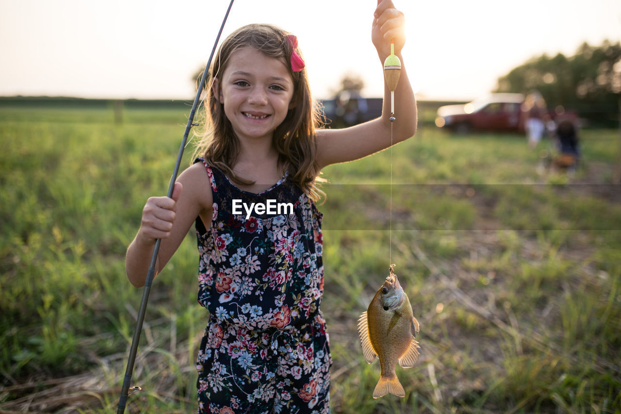 Portrait of smiling girl holding dead fish in fishing line on field during sunset
