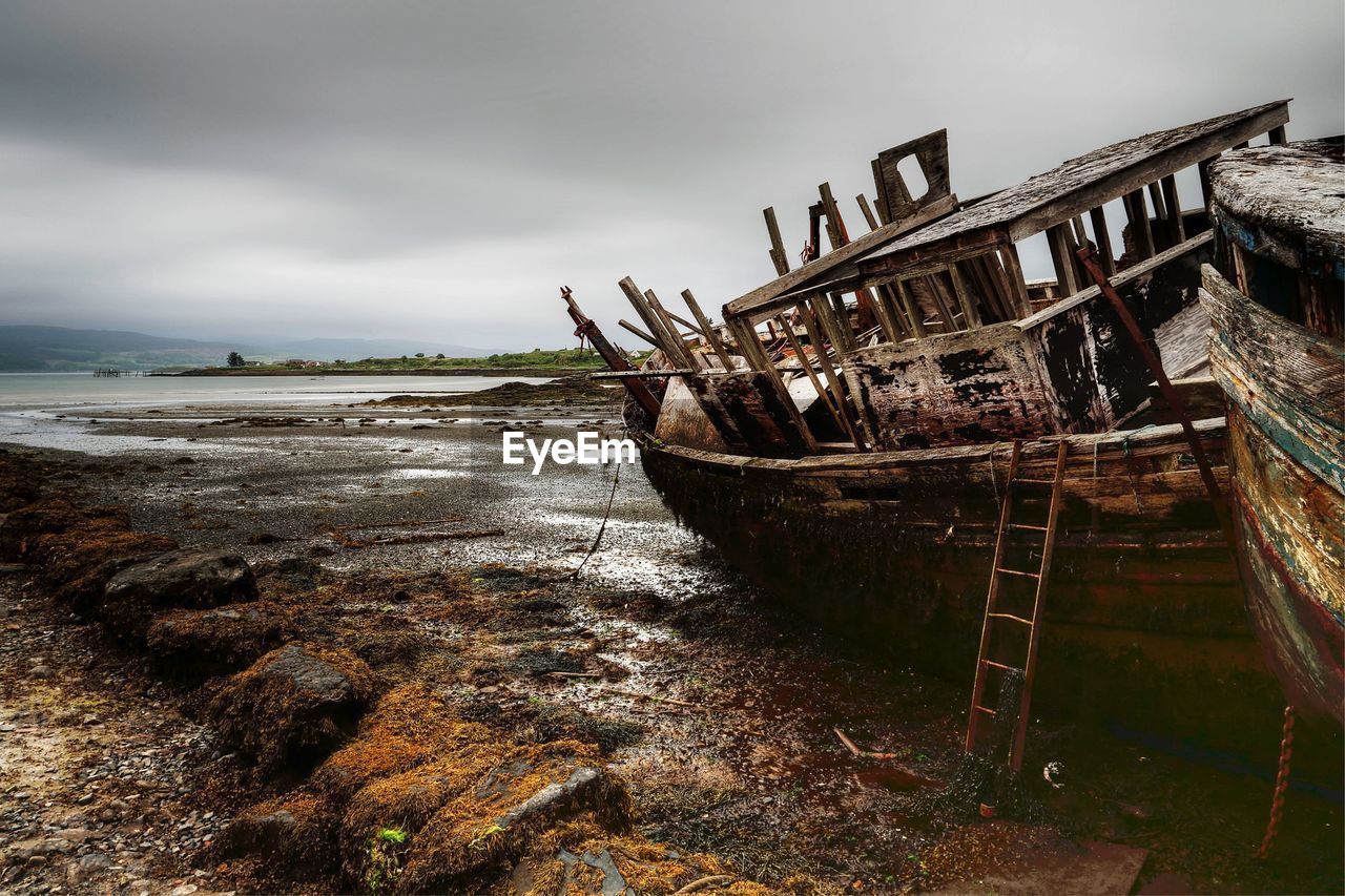 Broken abandoned boat moored at sea shore against cloudy sky