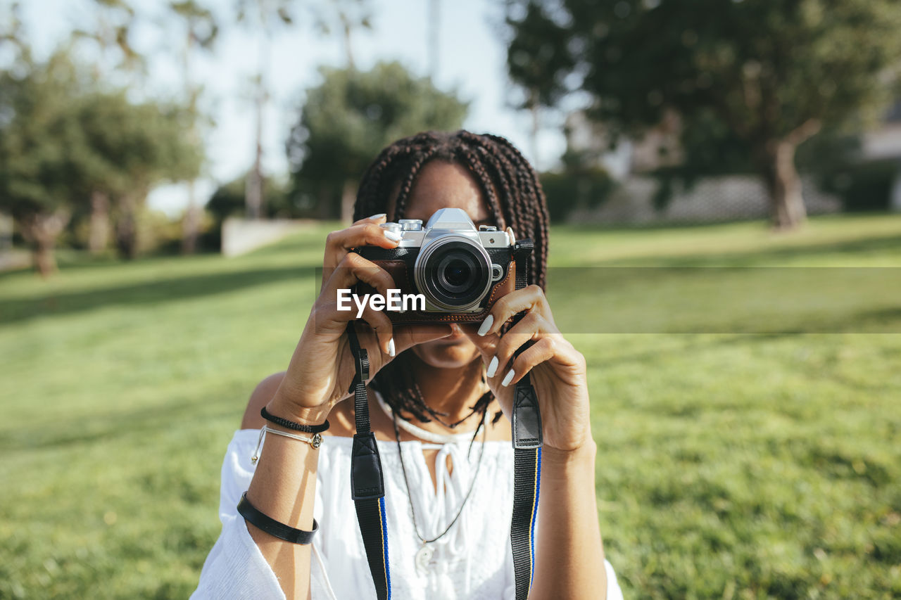 Unrecognizable african american female photographer taking picture and covering face with photo camera while standing on grassy lawn against blurred background