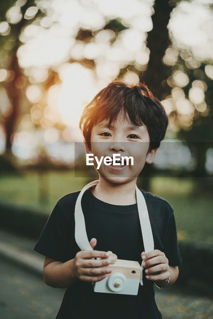 Portrait of happy boy with toy camera standing at park during sunset