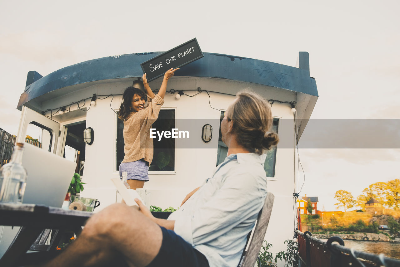 Boyfriend looking at girlfriend mounting placard at houseboat against sky