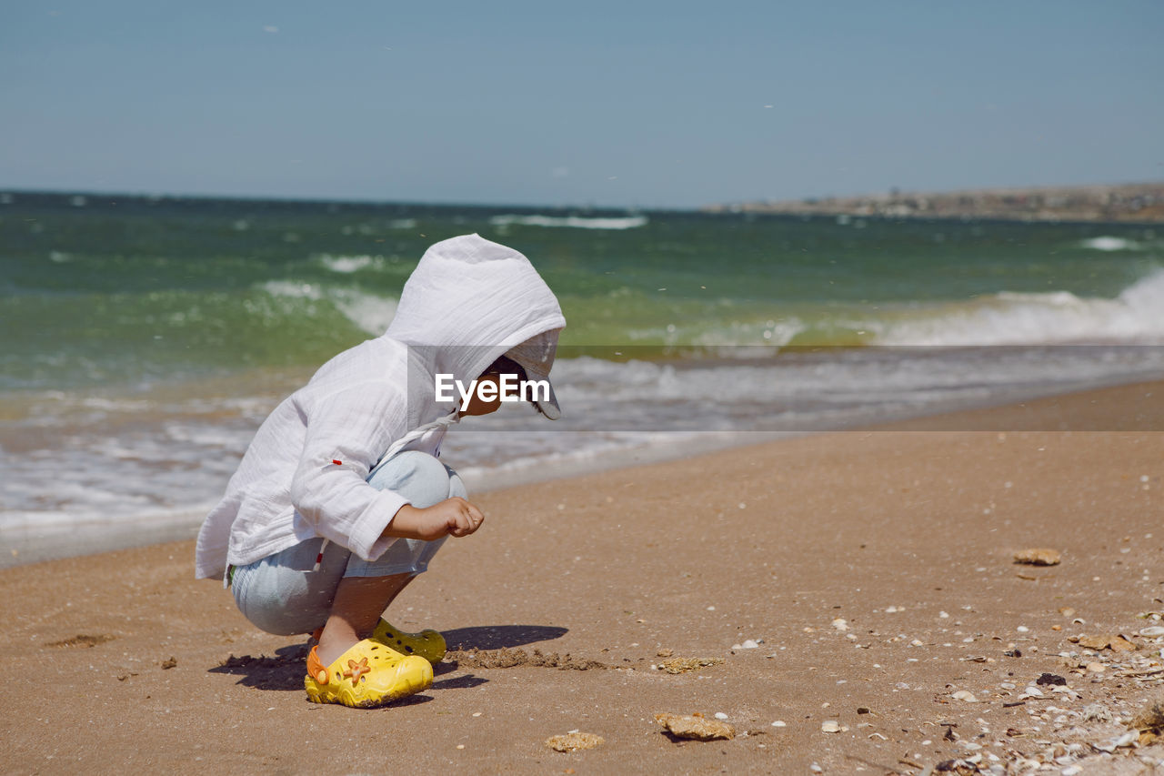 Baby boy sit on the beach with sand in yellow slippers and white shirt