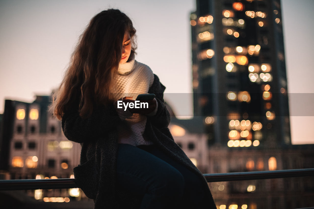 Teenage girl using smart phone sitting on railing in city at night