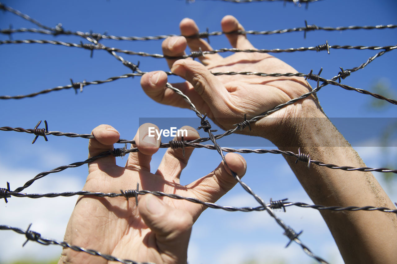 Cropped hands on barbed wire fence against sky