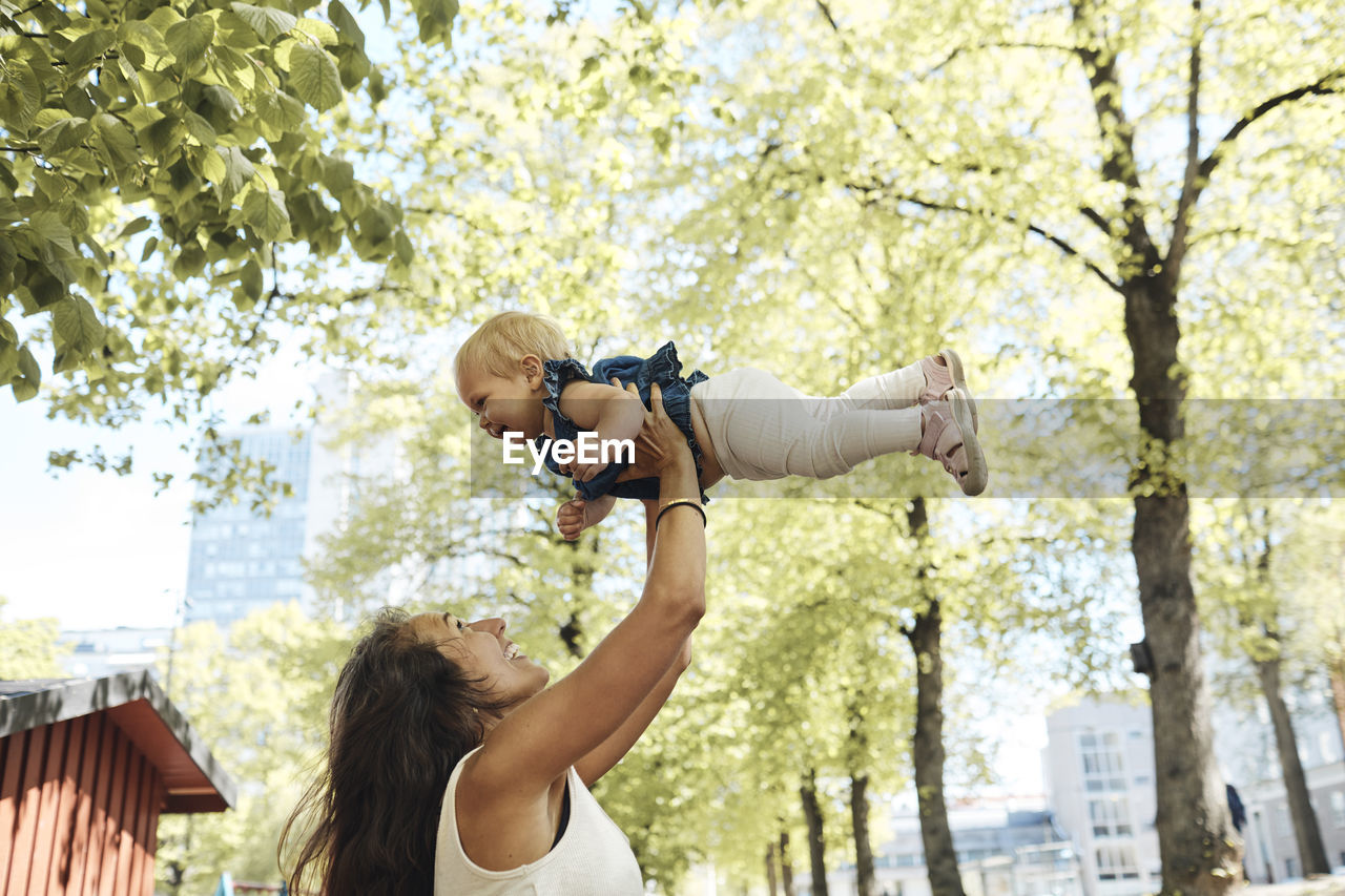 Side view of playful mother lifting daughter against trees