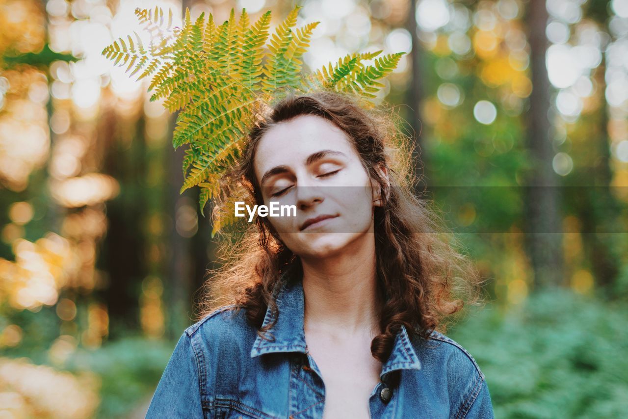 Eyes closed young woman with leaves on behind in forest