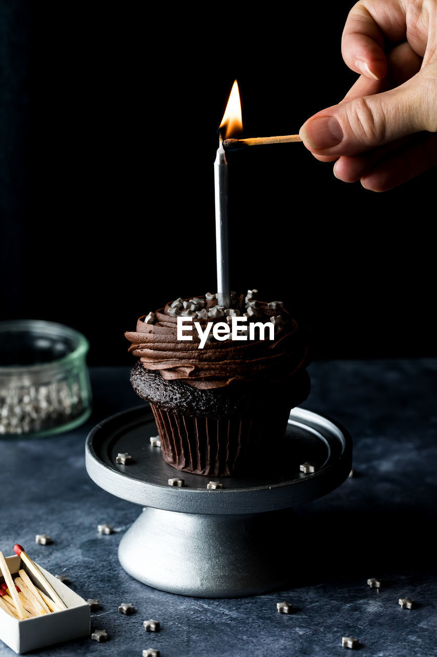 A hand lighting a candle on a chocolate cupcake, against a black background.