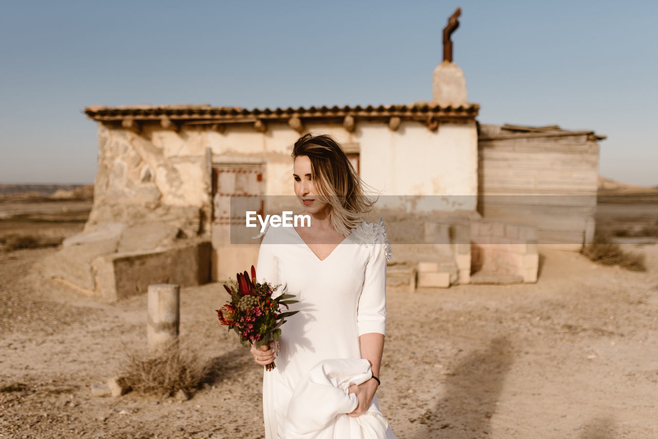 Woman in white dress and with flowers standing against shabby aged cabin on wedding day in bardenas reales natural park in navarra, spain