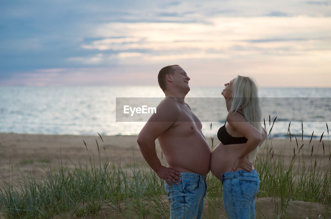 Young couple standing on beach against sky during sunset