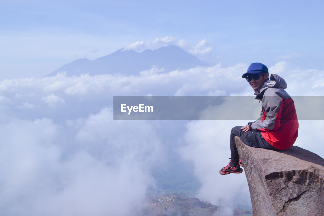 Portrait of man sitting on rock against cloudy sky