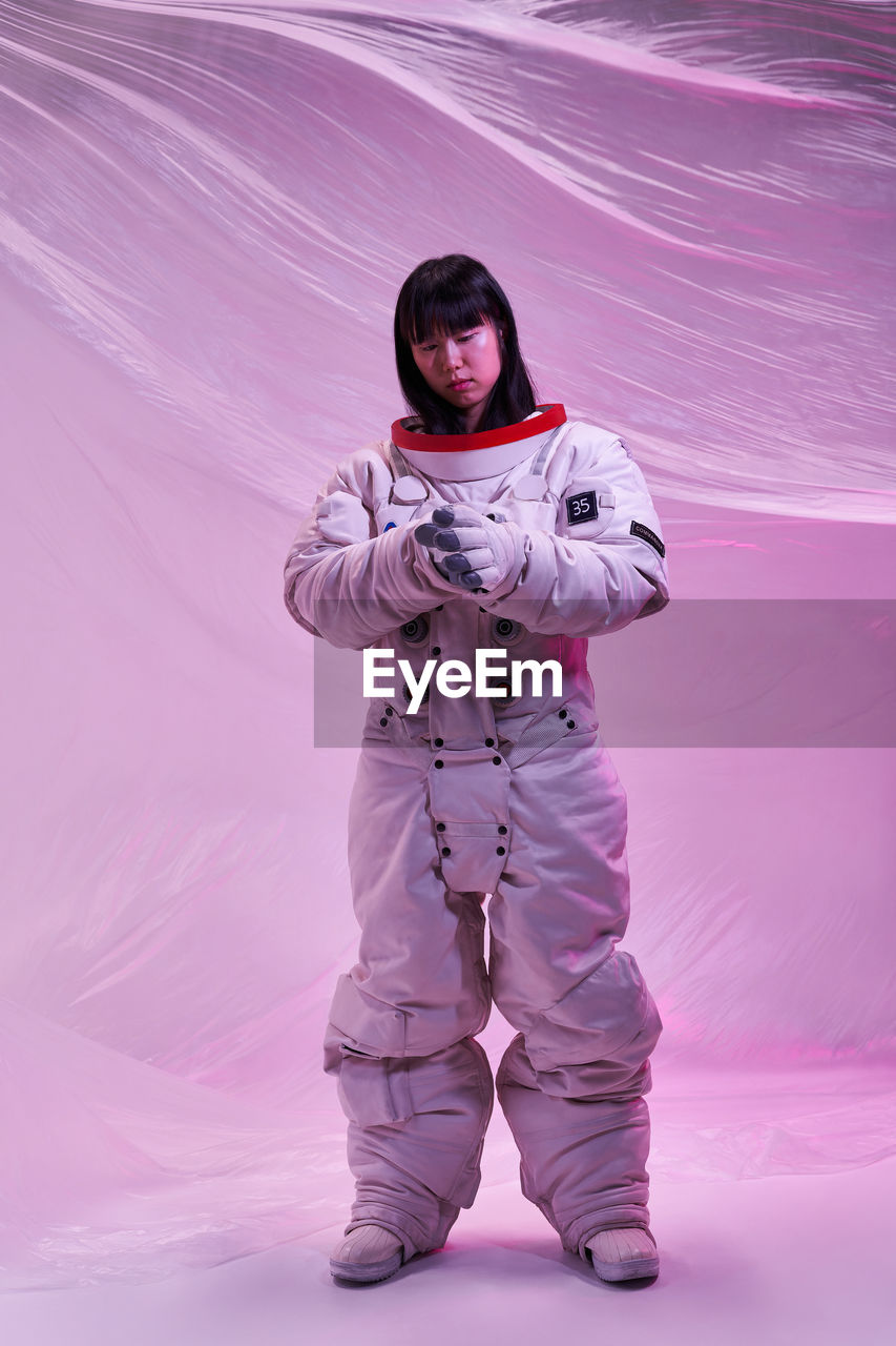 Full body asian woman in spacesuit adjusting gloves while standing against futuristic background under pink neon light