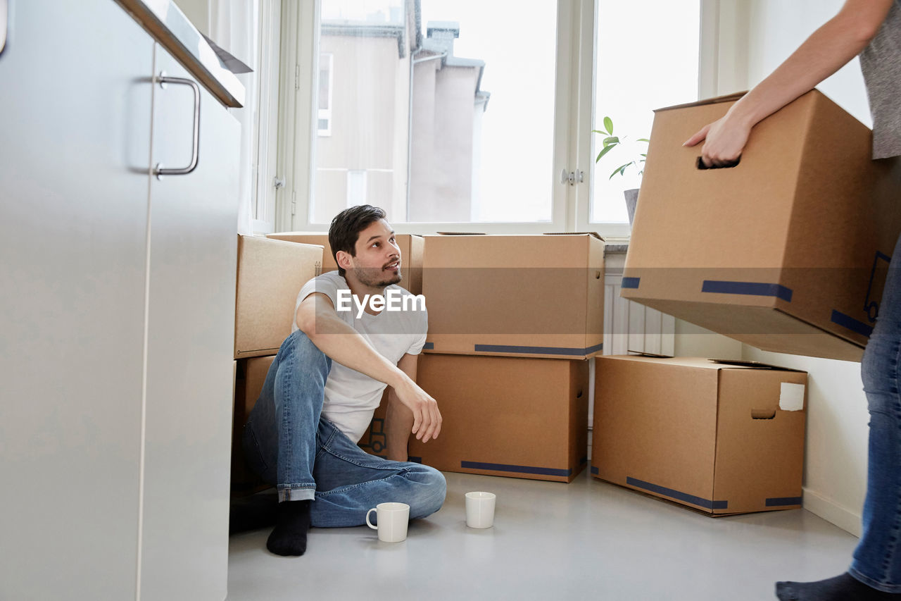 Man with coffee mugs looking at woman carrying cardboard boxes in new house