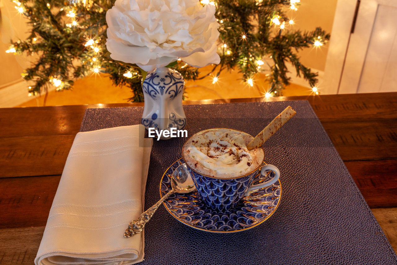 Cup of hot chocolate in a fancy bone china cup with gold and a silver spoon in front of a christmas