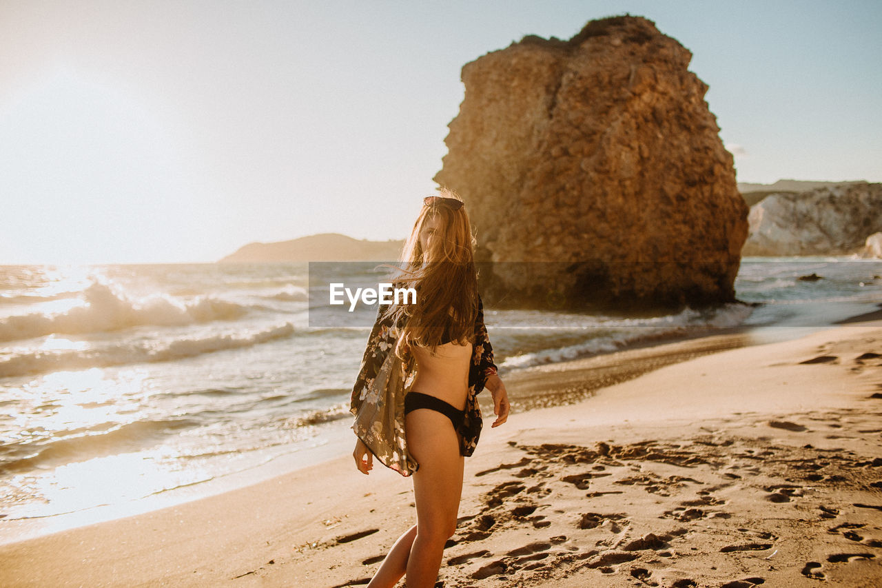 Side view slender young female in black swimwear with long hair standing on sandy seacoast near rough rocks on sunny hot day in fyriplaka milos