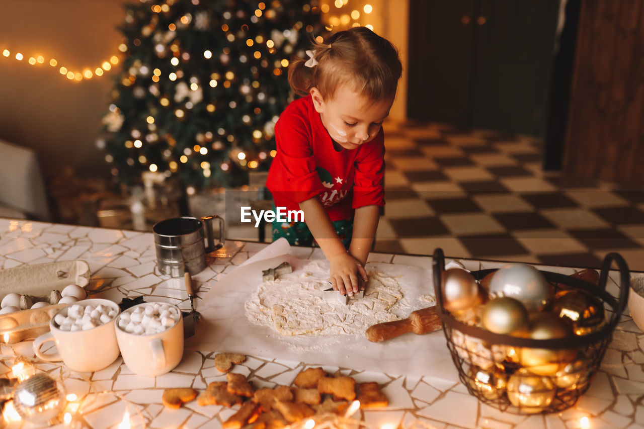 A little girl in red pajamas cooks and eats christmas cookies in the decorated kitchen of the house