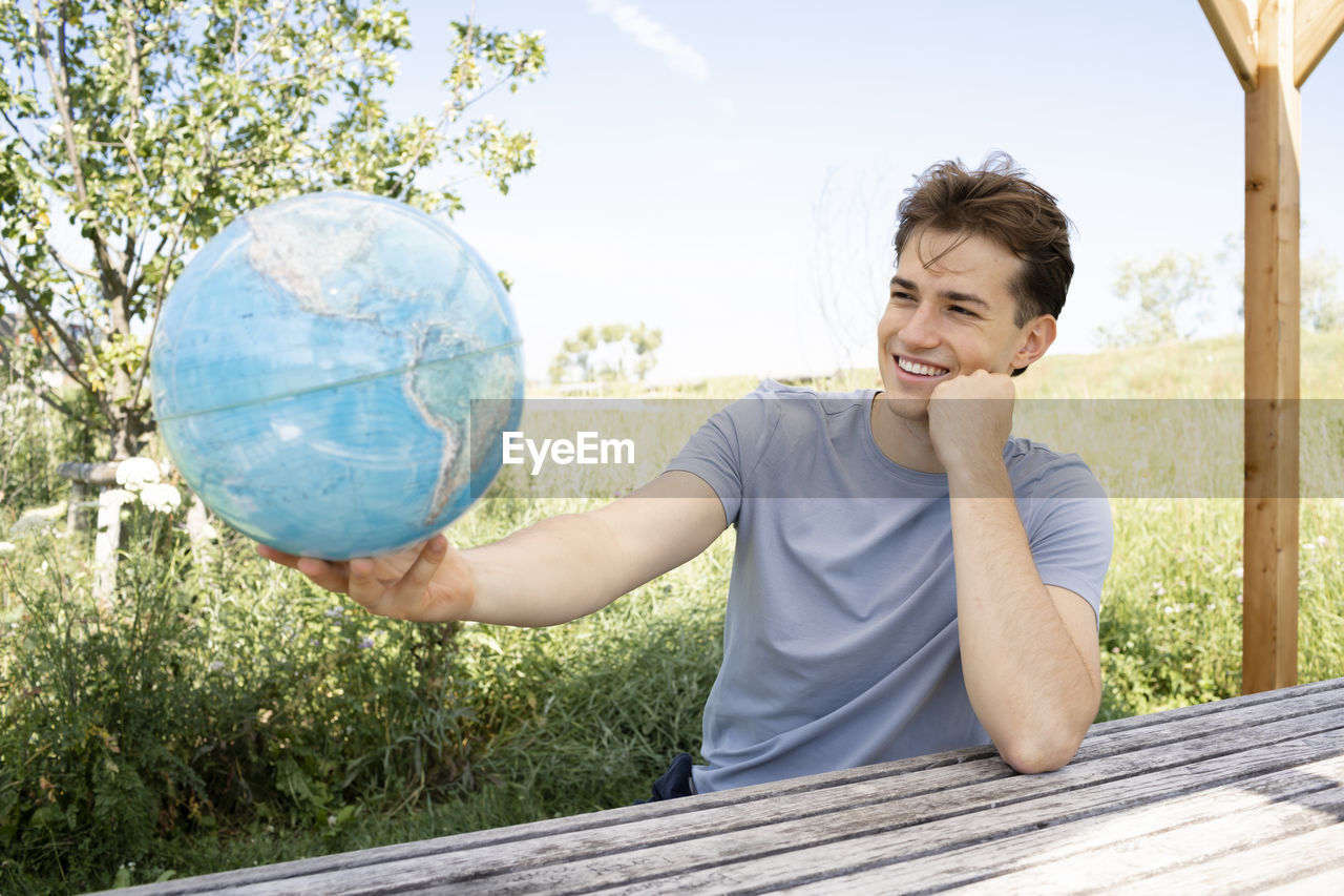 one person, nature, sky, smiling, casual clothing, happiness, day, globe - man made object, leisure activity, adult, outdoors, holding, front view, emotion, environment, tree, plant, environmental conservation, men, looking, vacation, portrait, sphere, lifestyles, child, person, cheerful, young adult, sunlight, enjoyment, ball, waist up, summer, childhood, spring, globe, relaxation, world, t-shirt