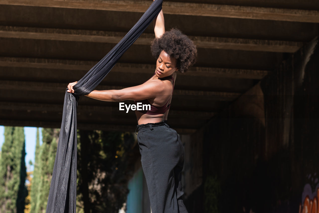 Slender african american female balancing on hanging hammock while doing aerial yoga above ground under bridge in city