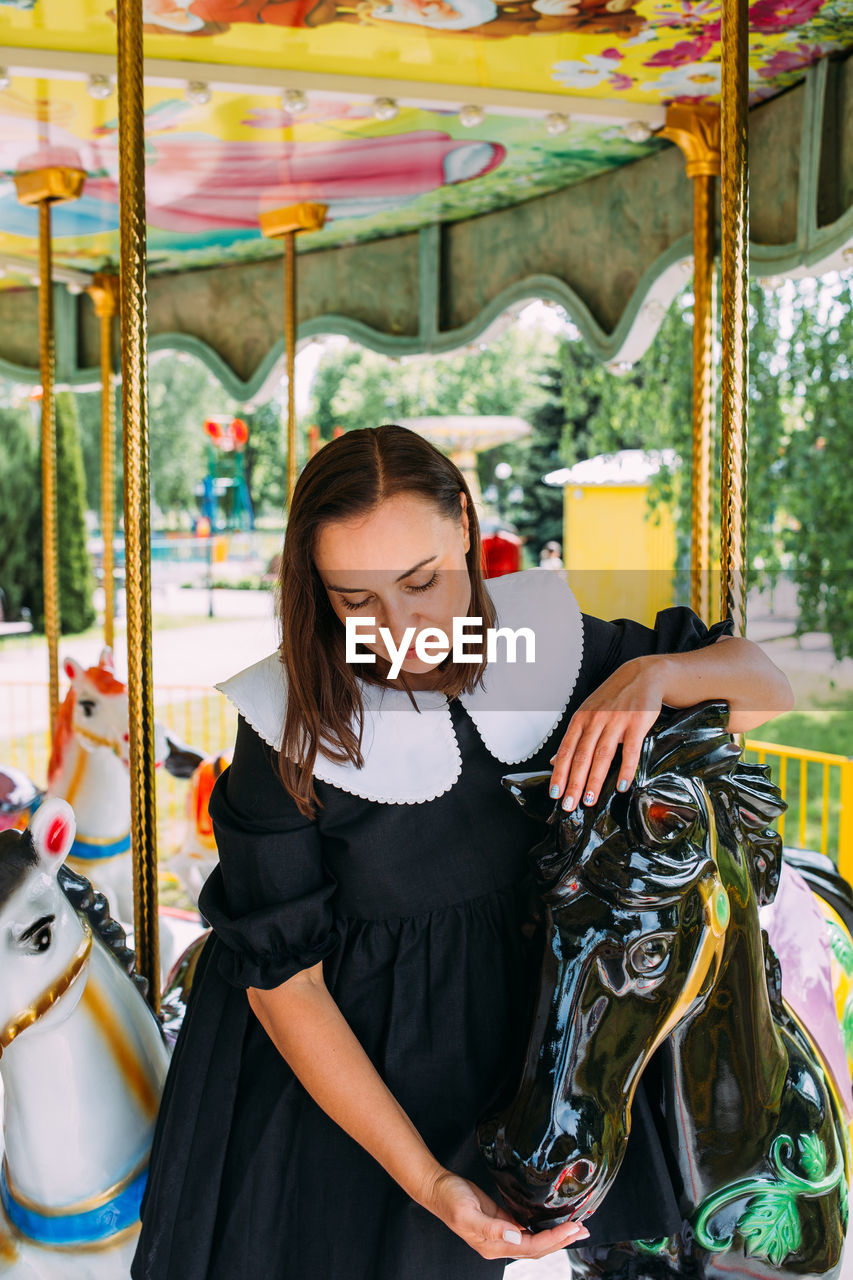 Beautiful brunette girl in a black dress poses on a bright carousel with horses