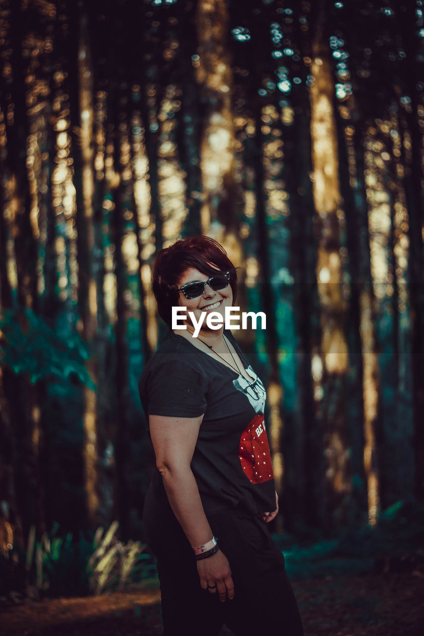 YOUNG WOMAN WEARING SUNGLASSES STANDING BY TREE TRUNKS