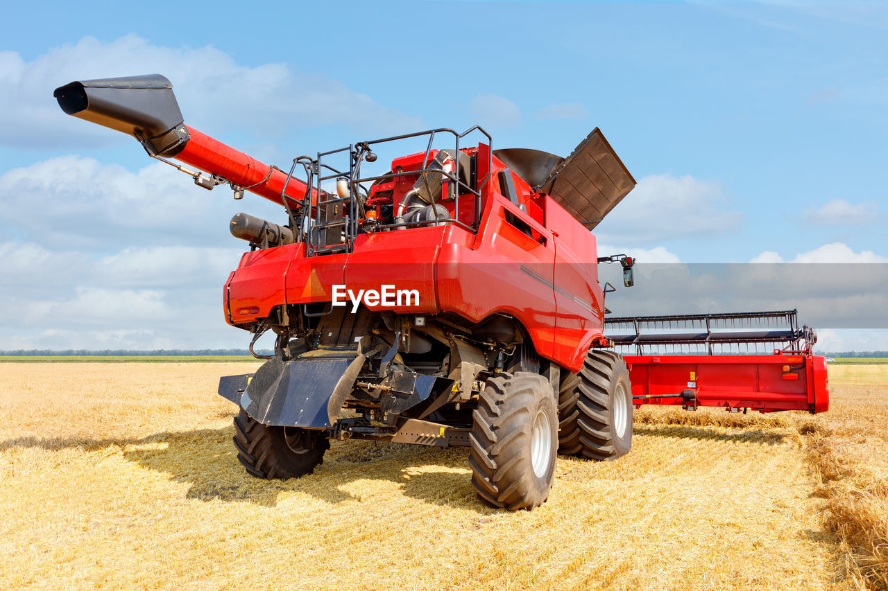 A large red agricultural harvester harvesting crops against the backdrop of a wheat field on  day.