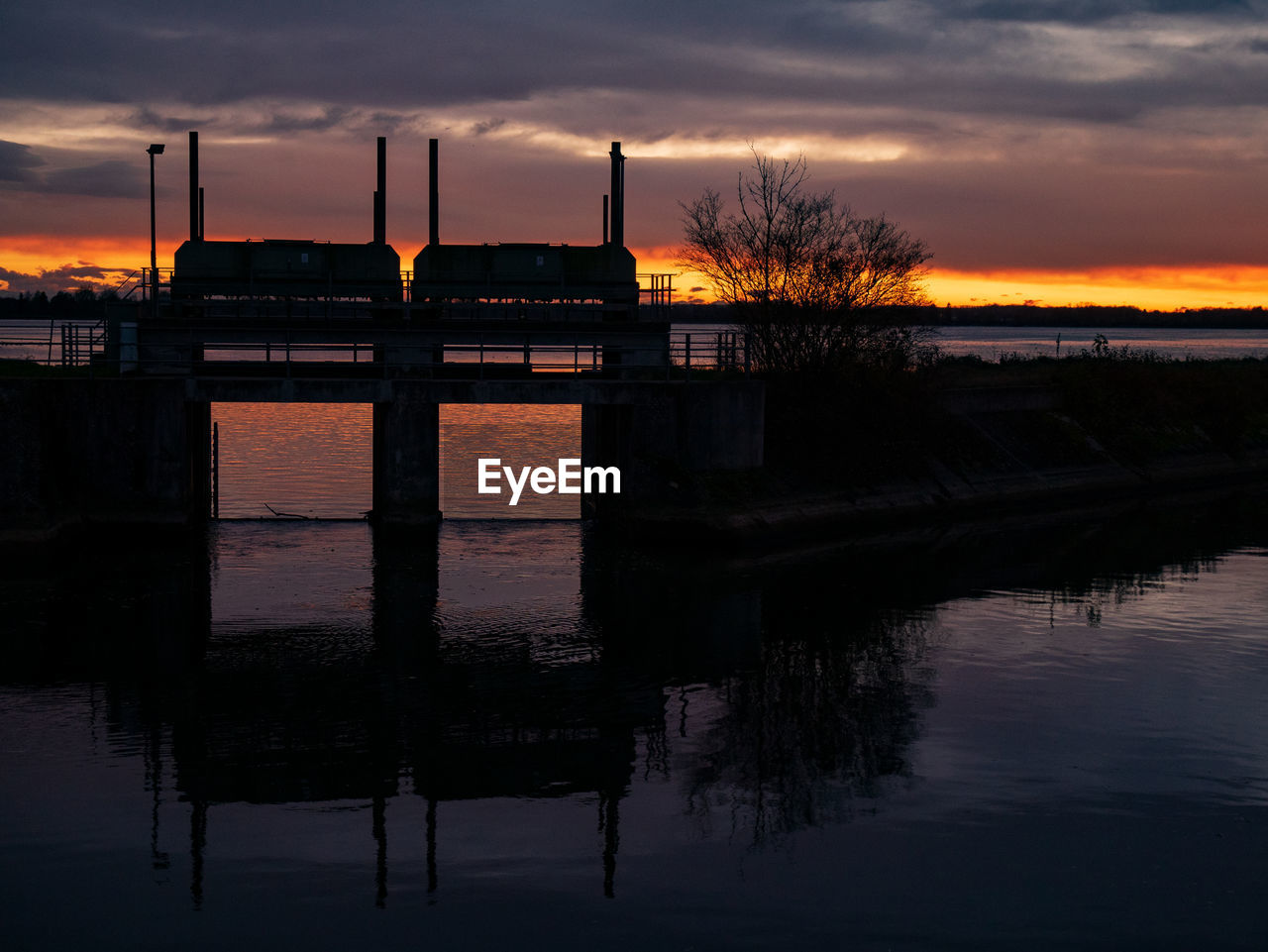 SILHOUETTE BUILT STRUCTURE BY LAKE AGAINST ORANGE SKY