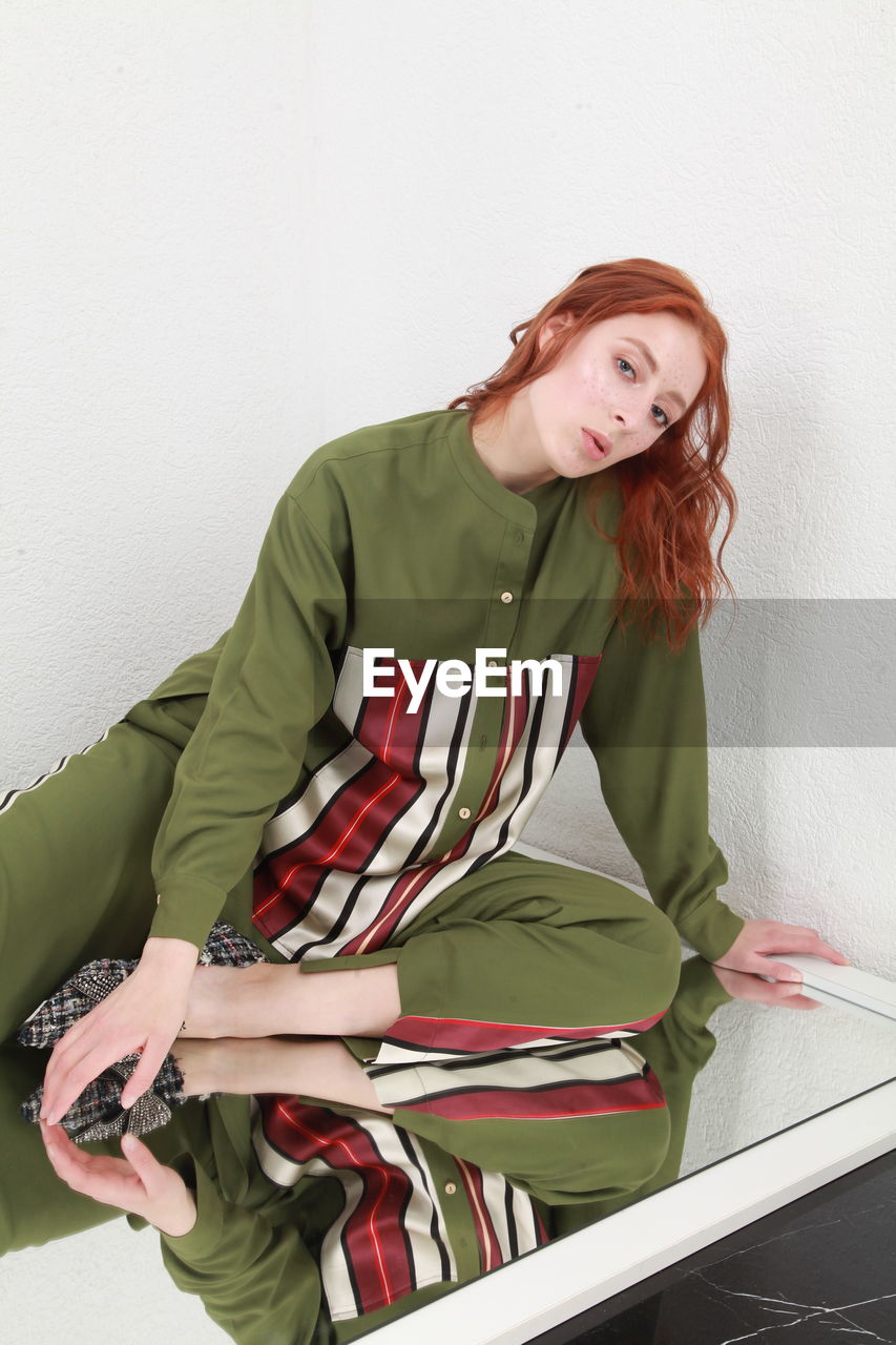 Cropped portrait of woman sitting on huge mirror in green stylish outfit against white wall studio