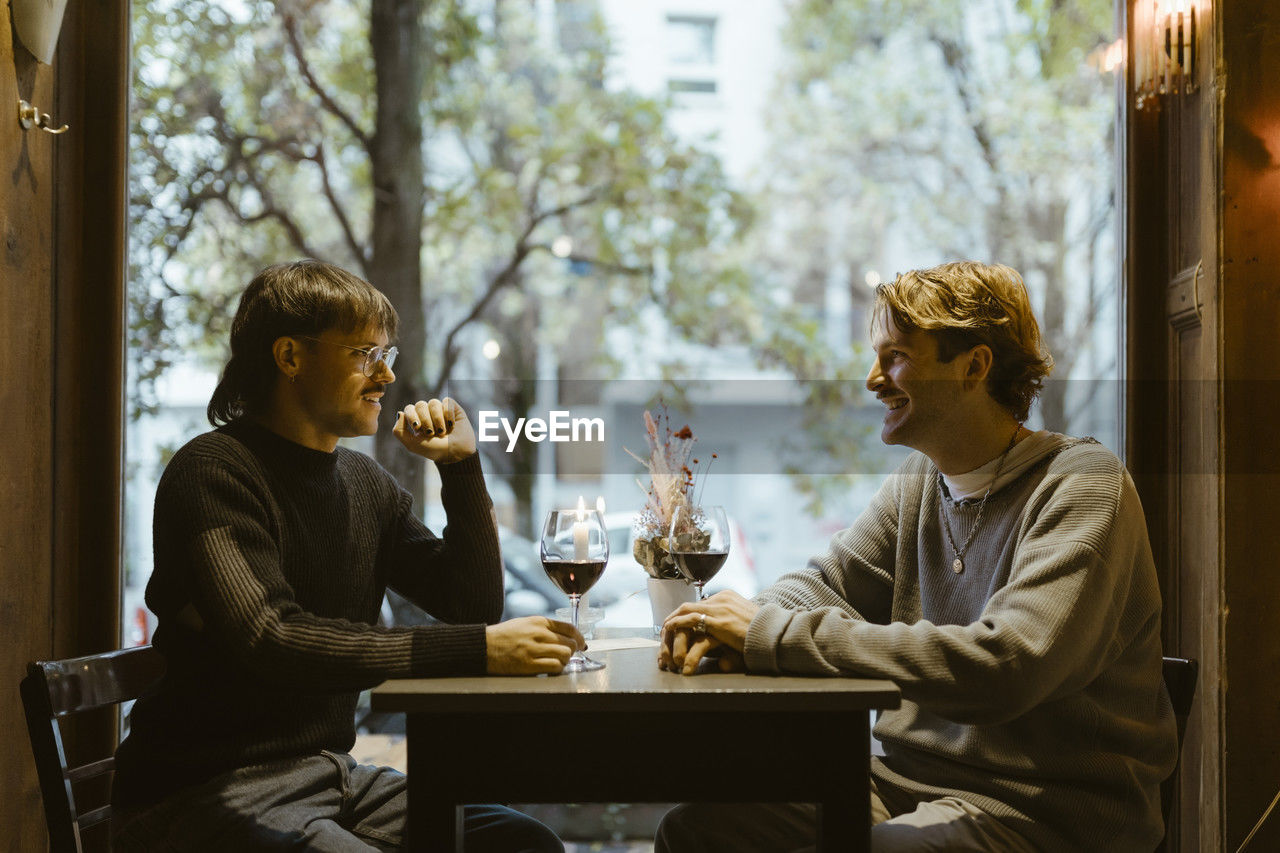 Happy gay couple talking while sitting with wineglasses on table during date at restaurant