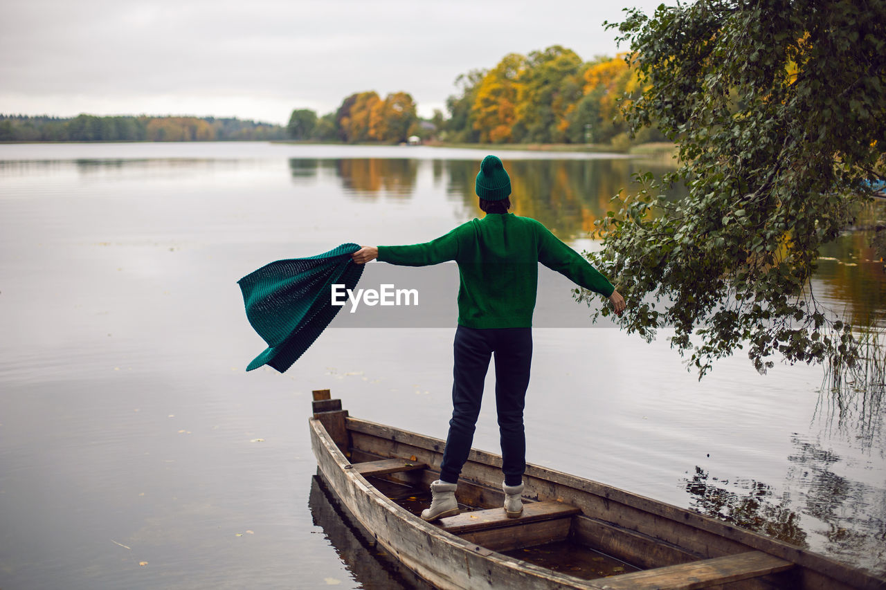 Woman in a green sweater stands with her back in an old wooden boat on a lake in autumn