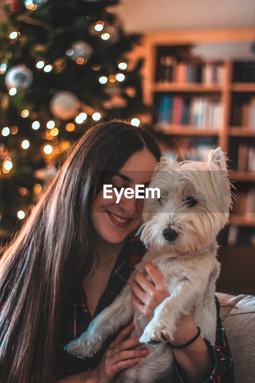 Smiling woman with dog against christmas tree at home