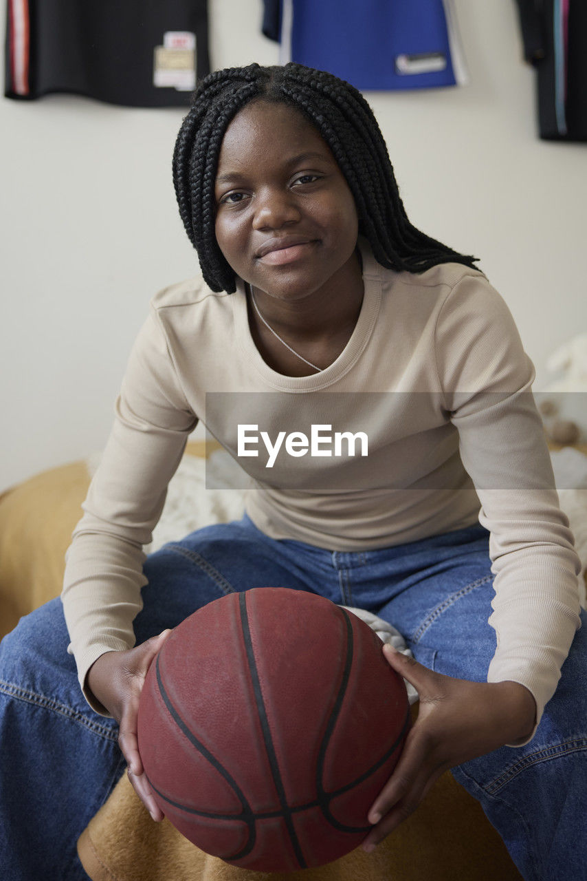 Portrait of teenage girl holding basketball while sitting in bedroom at home