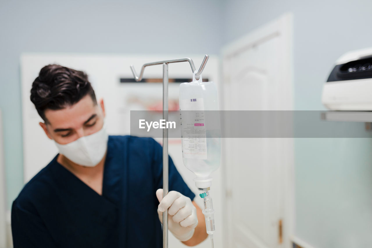 Anonymous young male nurse in respiratory mask installing drop counter with solution in transparent bag in hospital