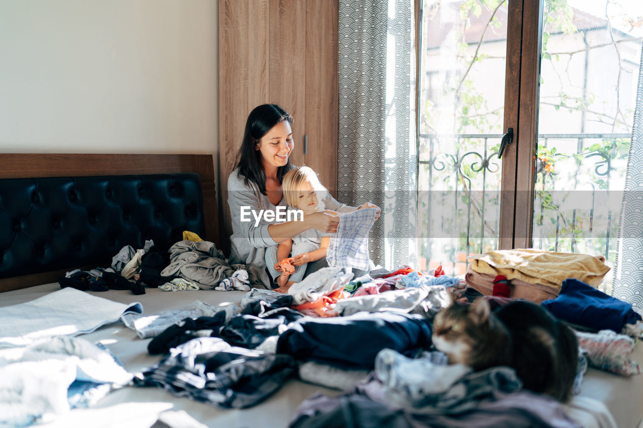 Mother doing chores while sitting with daughter on bed