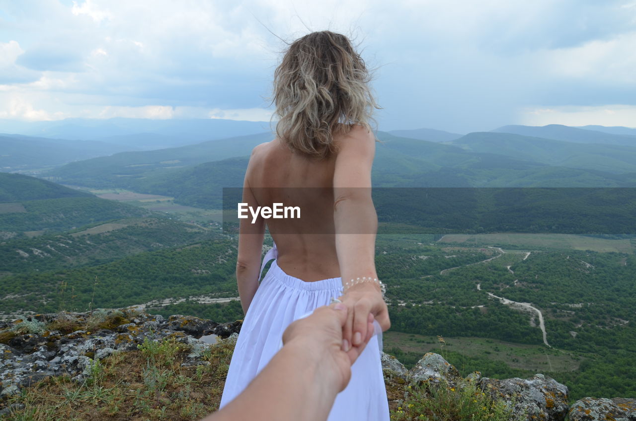 Midsection of shirtless woman looking at mountains against sky