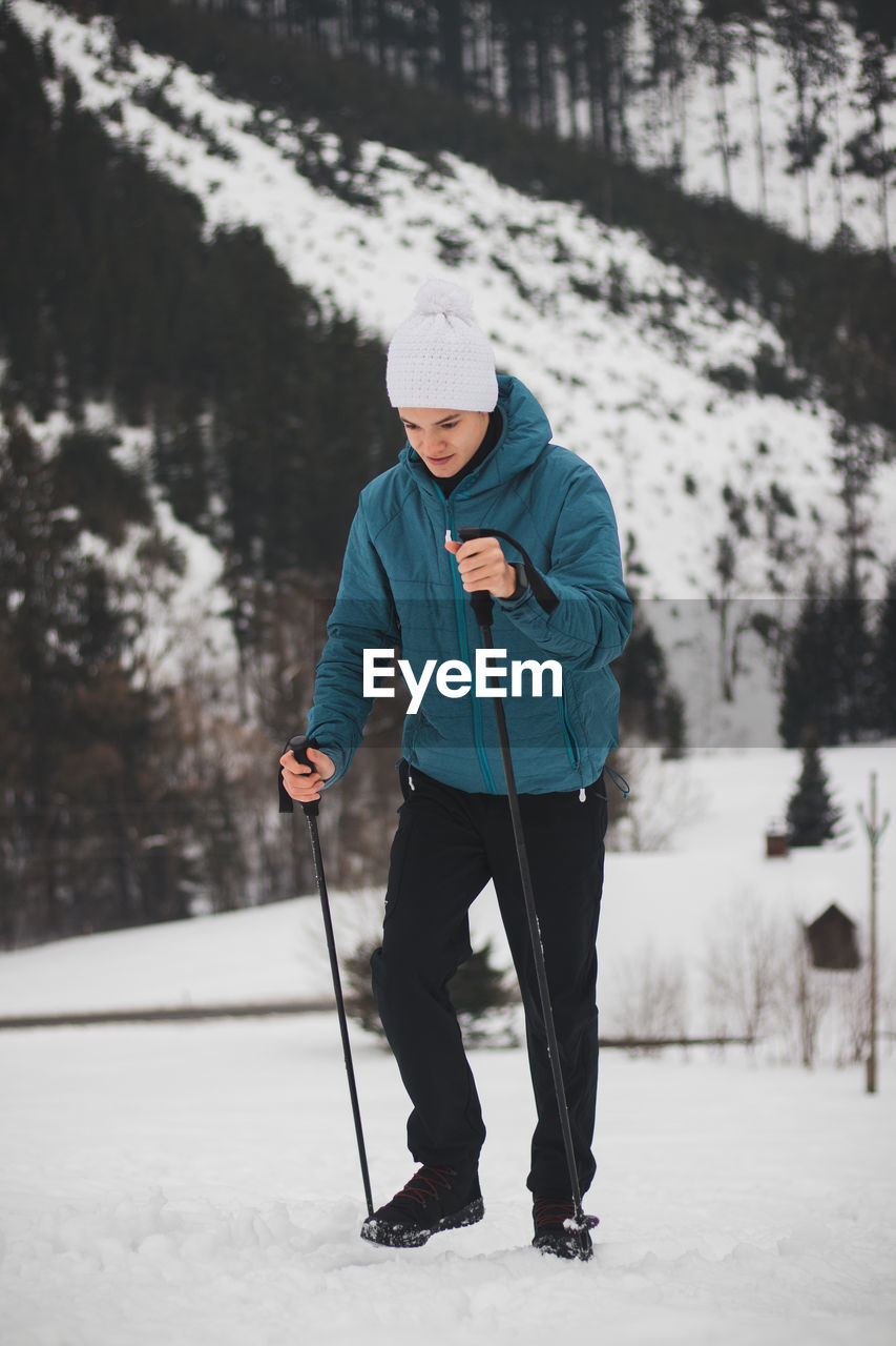 Teenager in a white cap with a pompom and a blue jacket climbs a mountain using trekking poles 