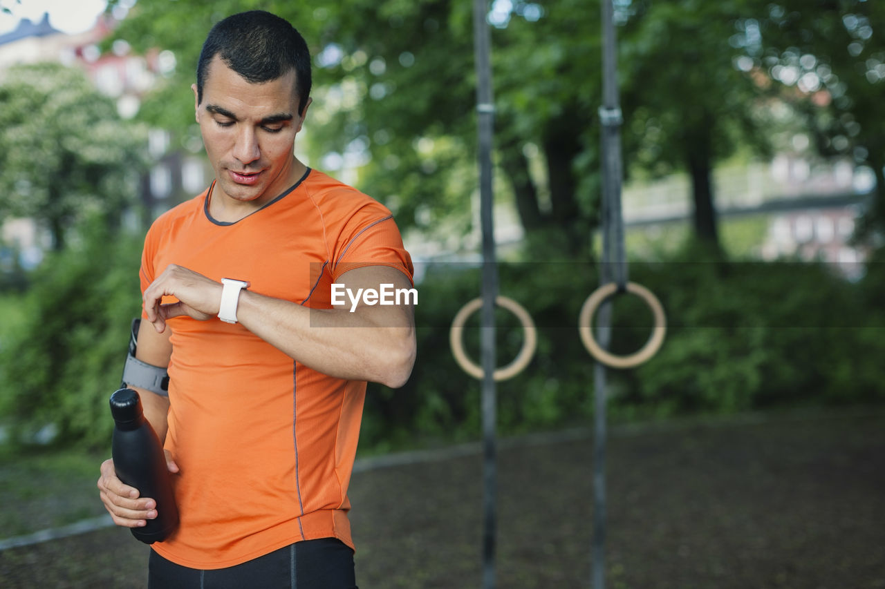 Man checking smart watch and holding bottle while standing at park