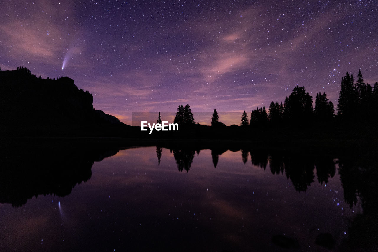 Reflection of silhouette trees in lake against sky at night