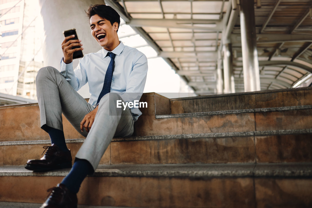 FULL LENGTH OF YOUNG MAN USING MOBILE PHONE WHILE SITTING ON FLOOR