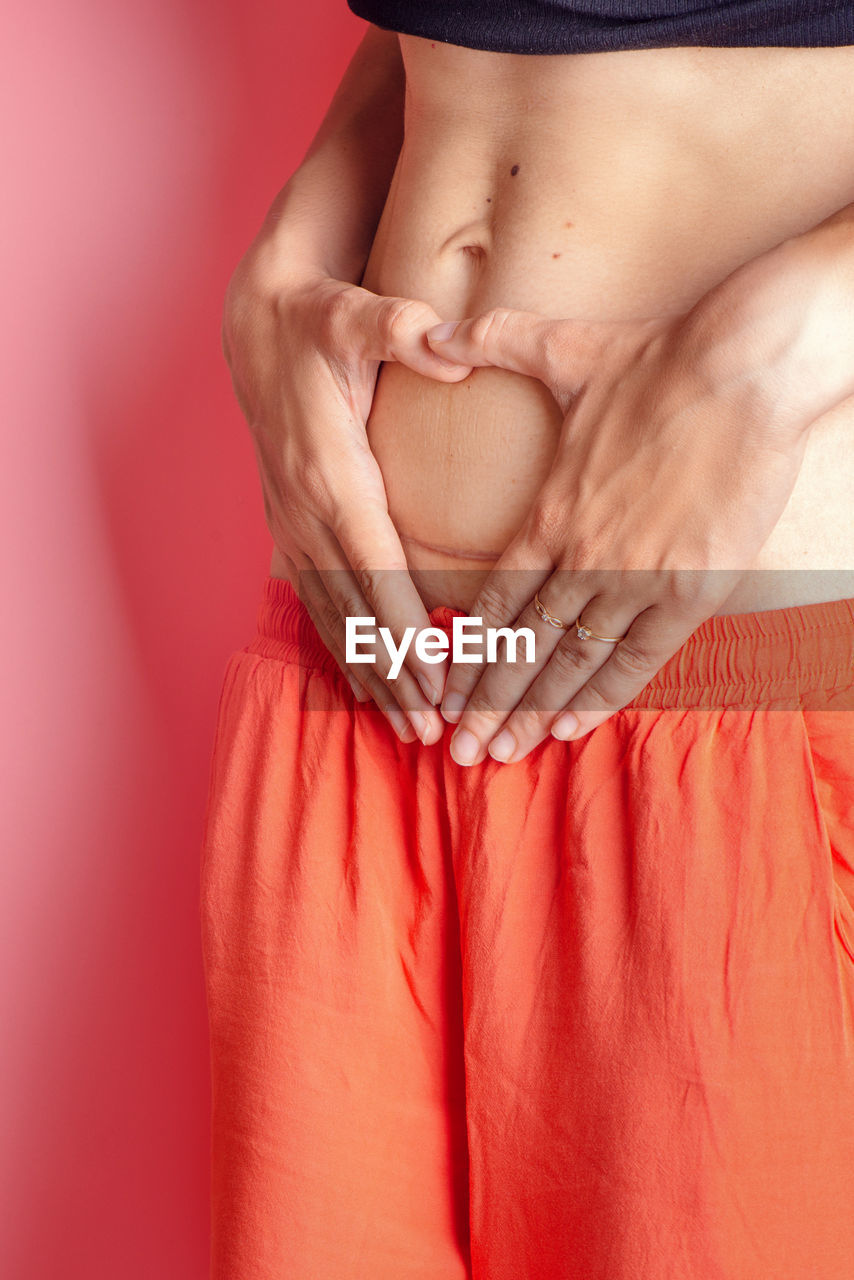 Midsection of woman making heart shape on belly against pink background