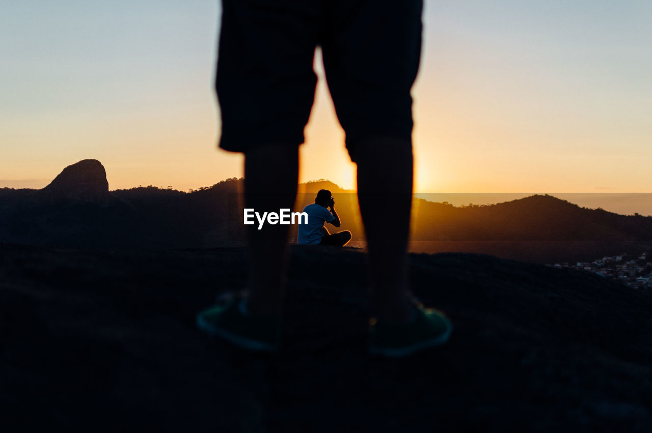 Low section of man standing on rock against photographer during sunset