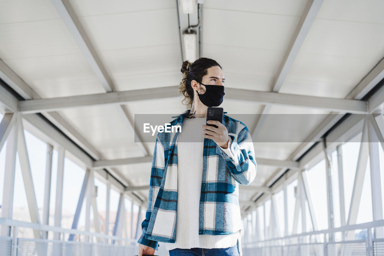 Young man wearing face mask looking away while using mobile phone standing on bridge