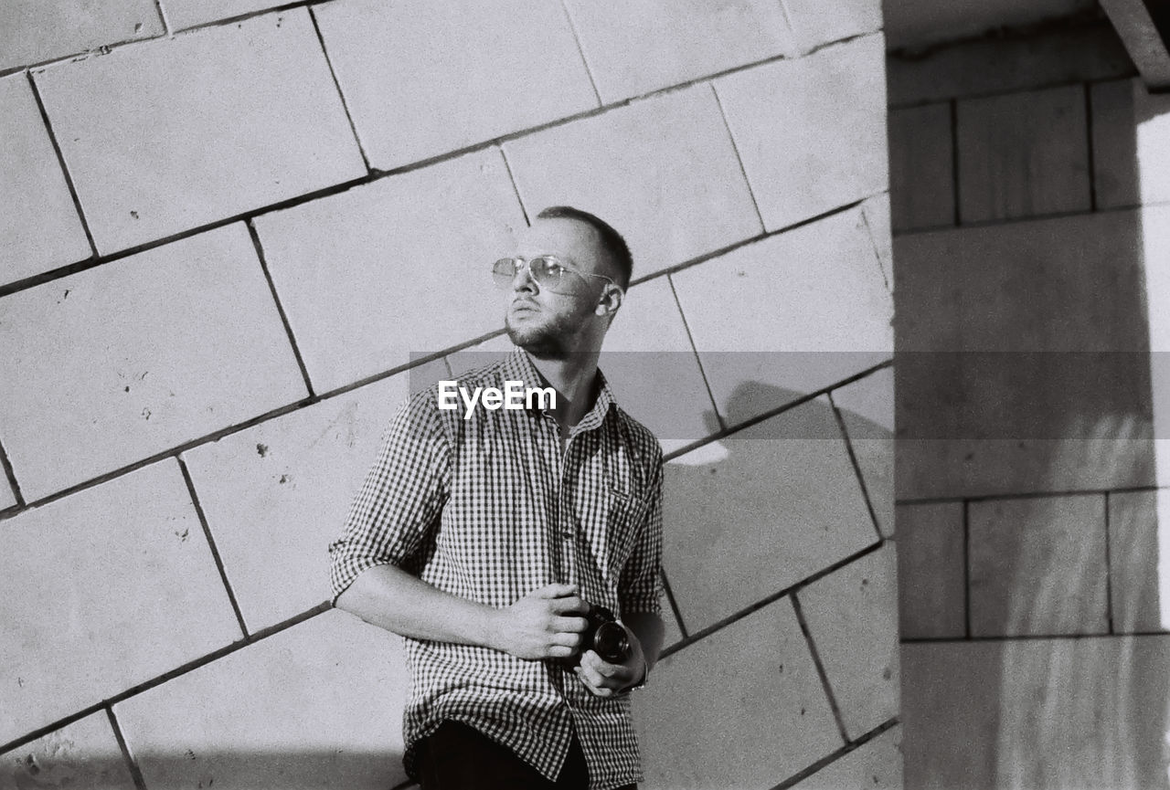 Young man wearing sunglasses looking away while standing against wall