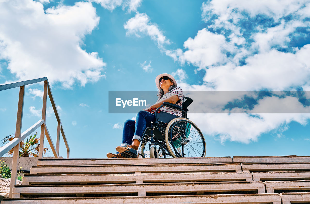 Low angle full body of positive handicapped female sitting in wheelchair near stairway looking away against blue sky in city