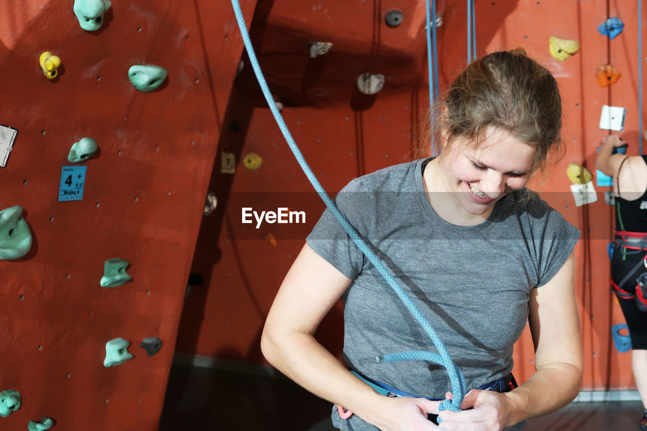 Smiling woman tying rope while standing against climbing wall