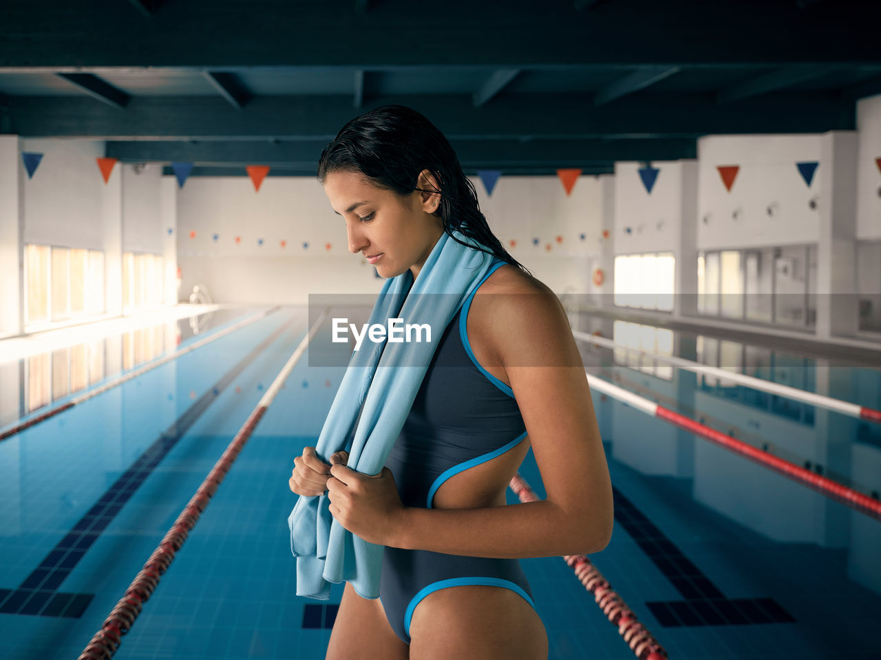 Side view of young female athlete in swimwear with towel and wet hair looking down against swimming pool after training