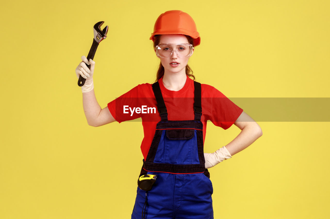 Portrait of young woman holding wrench against yellow background