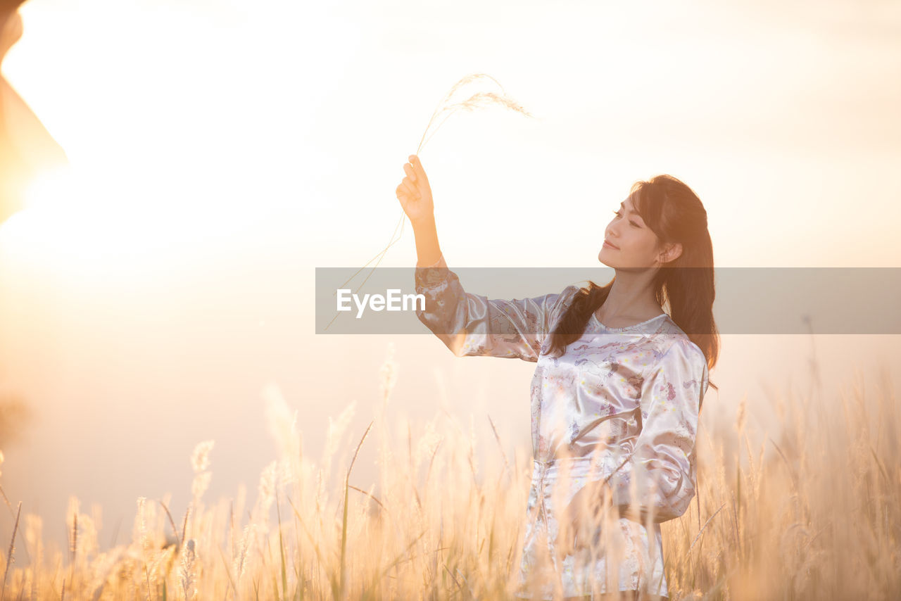 Mid adult woman holding plant while standing on field against sky during sunset