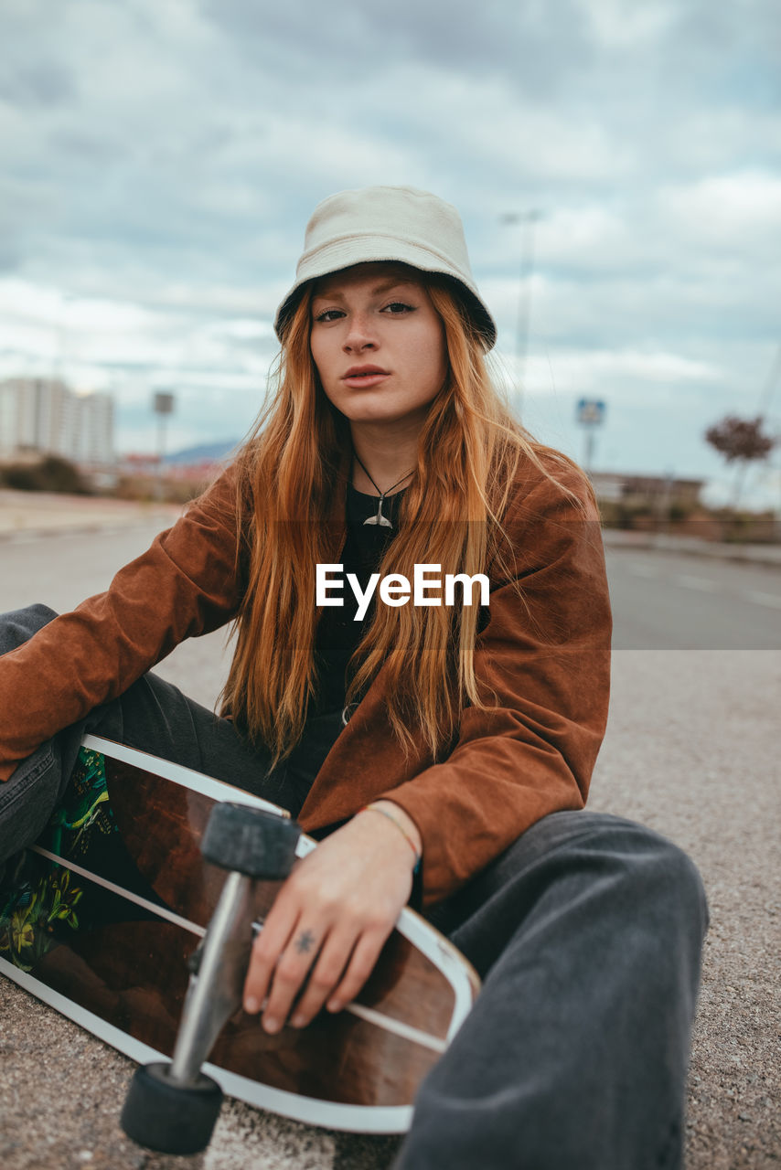 Young female skater with long blond hair in trendy outfit sitting on asphalt road with cruiser skateboard in hand against cloudy sky in countryside